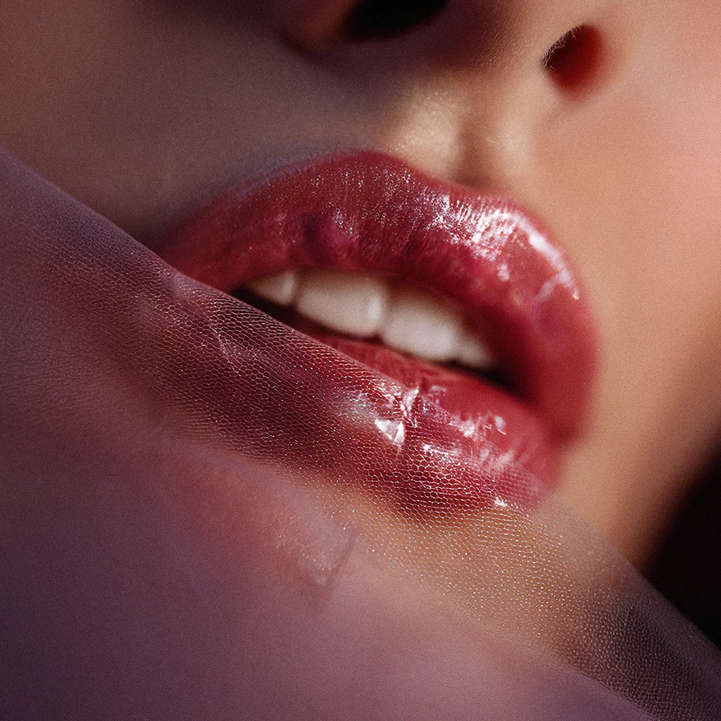 Nahaufnahme von sinnlichen, leicht geöffneten Lippen mit glänzendem, rötlichem Lipgloss, die teilweise von einem feinen, violetten Schleier verdeckt sind