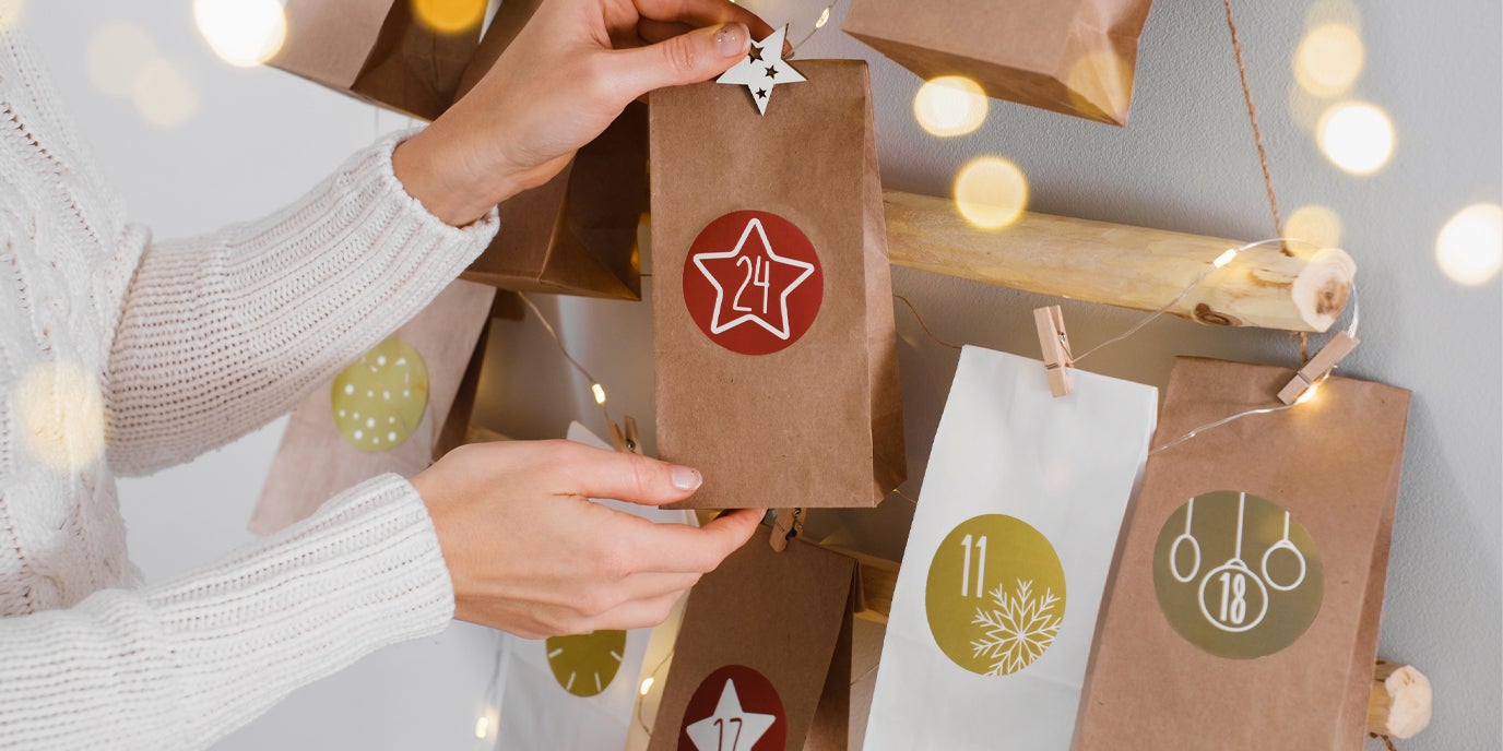 Hände hängen eine braune Papiertüte mit einem roten Stern und der Zahl 24 an einen selbstgemachten Adventskalender; weitere Tüten mit Zahlen und weihnachtlichen Motiven sind an einer Holzstange befestigt.