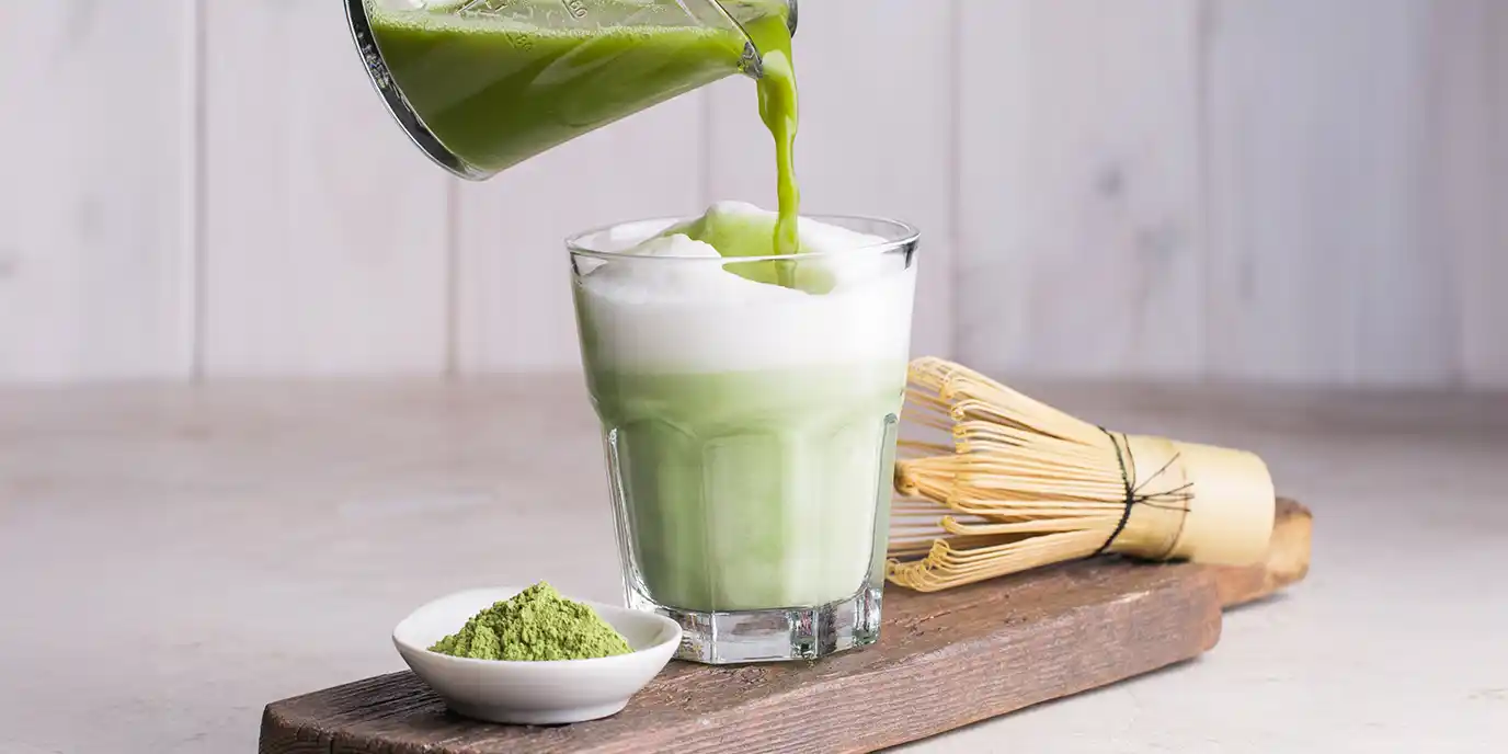 Ein Glas mit schaumigem Matcha Latte wird mit grünem Matcha-Tee aus einem Messbecher aufgegossen. Daneben stehen ein Bambusbesen und eine kleine Schale mit Matcha-Pulver auf einem Holzbrett.