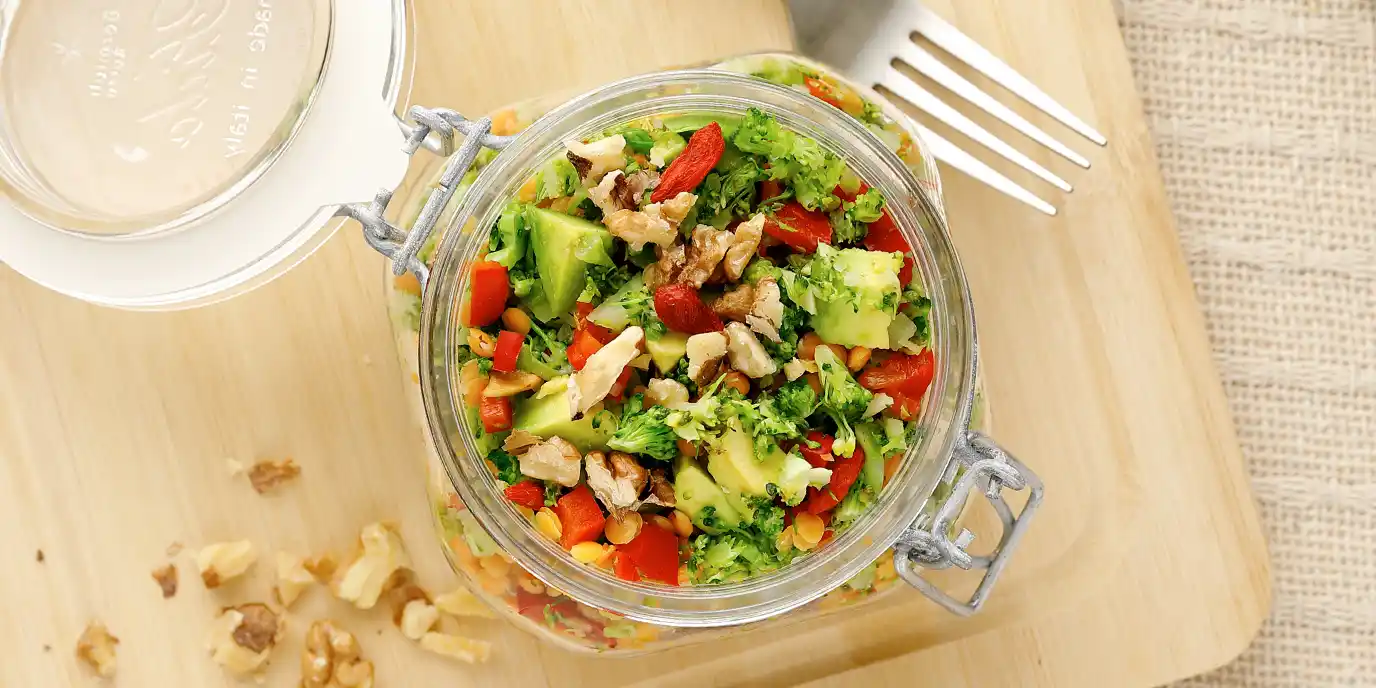 Bunter Salat mit Avocado und Nüssen in einem Einmachglas auf einem Holzbrett, daneben eine Gabel und ein paar Walnüsse.